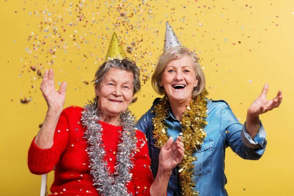 two middle aged women in party hats and leis toss confetti to celebrate hard working women who enjoy life