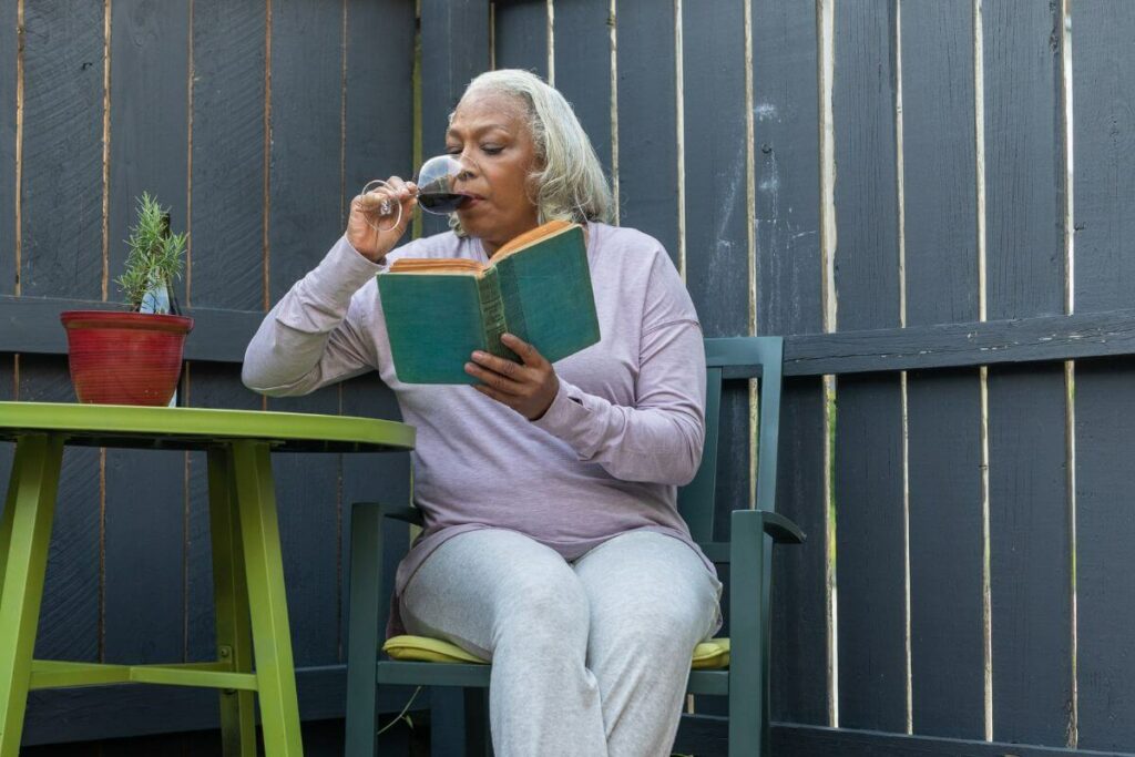 middle aged woman sits outside in her yard having a glass of wine and reading a book for a self care weekend