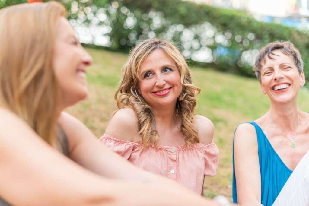 three middle aged women sit outside in a park laughing together for self care