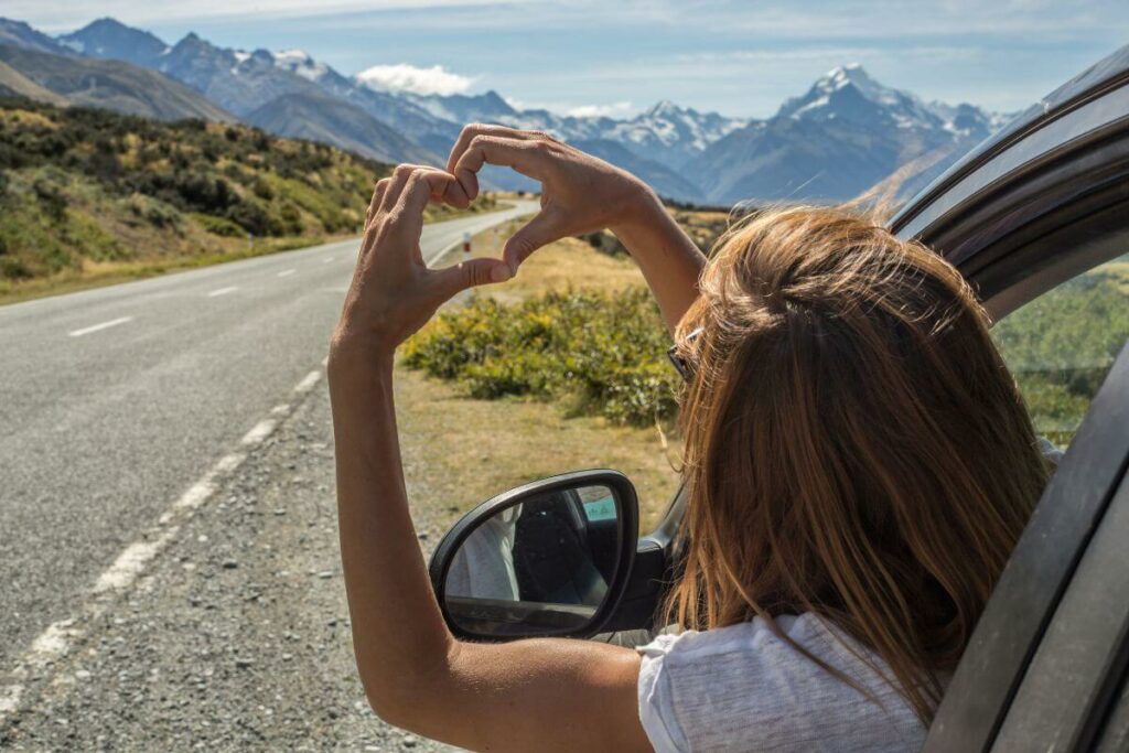 woman reaching out of a car window making a heart with her hands as she sits on the side of the road in the mountains