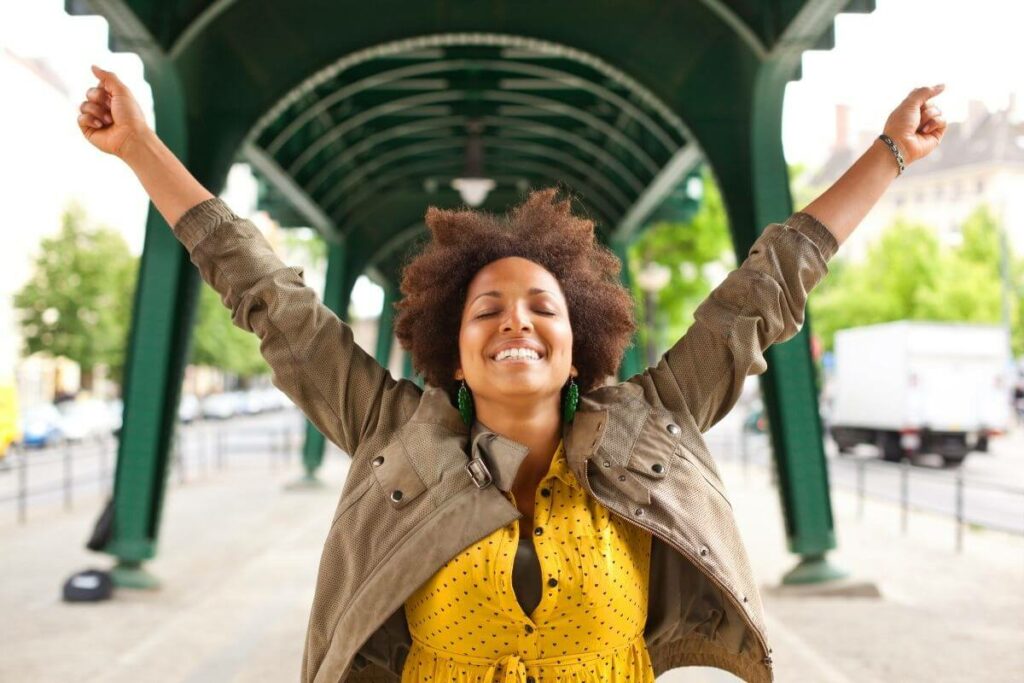 beautiful black woman happily throws her arms up in the air aftter reading powerful quotes reminding her to talk to yourself like someone you love