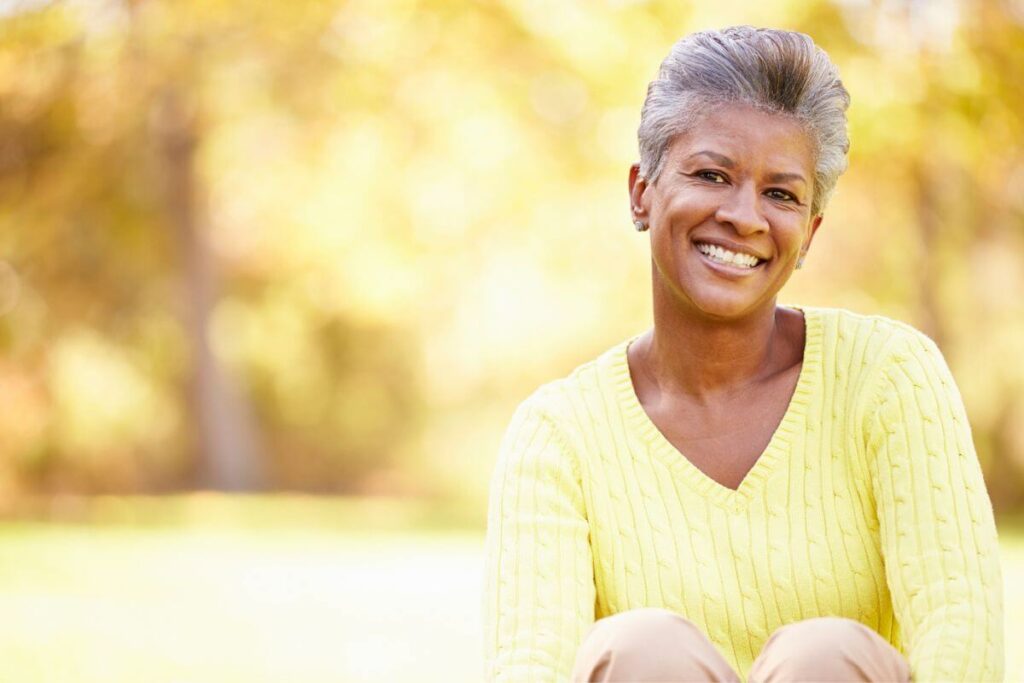 a dark skinned middle aged woman wearing a yellow sweater confidently sits in a park smiling