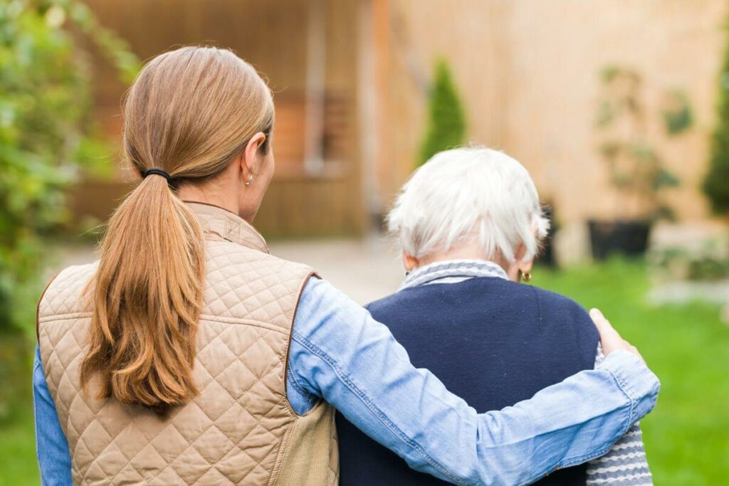 the backs of an adult child with her arm around her elderly mom walking in the park