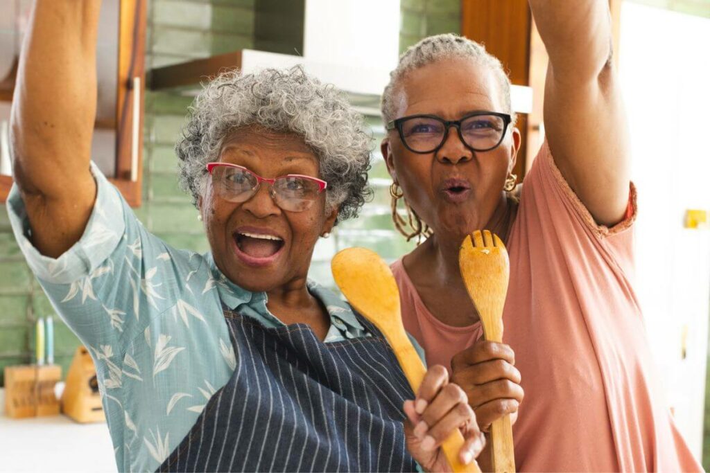 two elderly ladies pretend to sing into their wooden spoons while baking