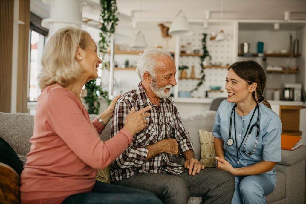 elderly wife and a paid professional caregiver sit with the elderly husband and talk about happy memories.