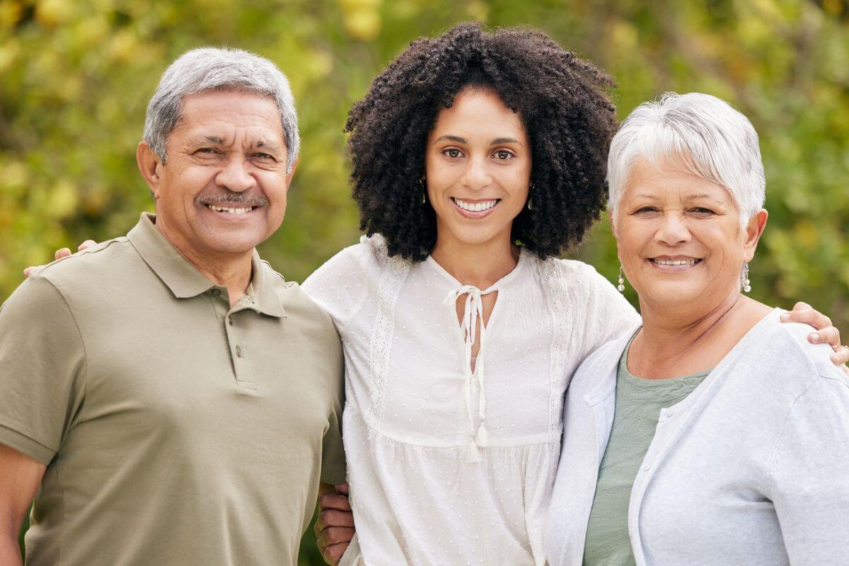 adult daughter embraces her elderly mother and father for a photo in the park.