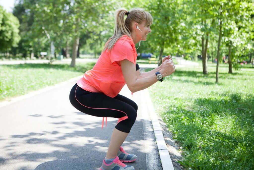 happy middle aged woman on a walk stops to do squats as part of her self-care Sunday routine.