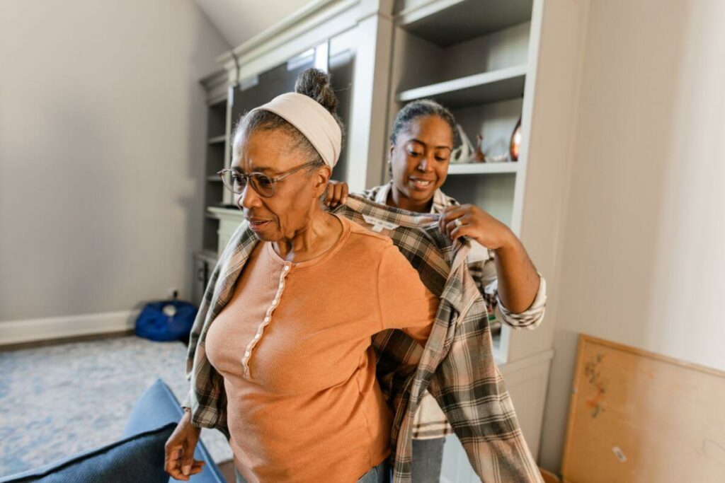 a middle aged daughter helps her elderly mother get dressed in adaptive clothing for seniors.
