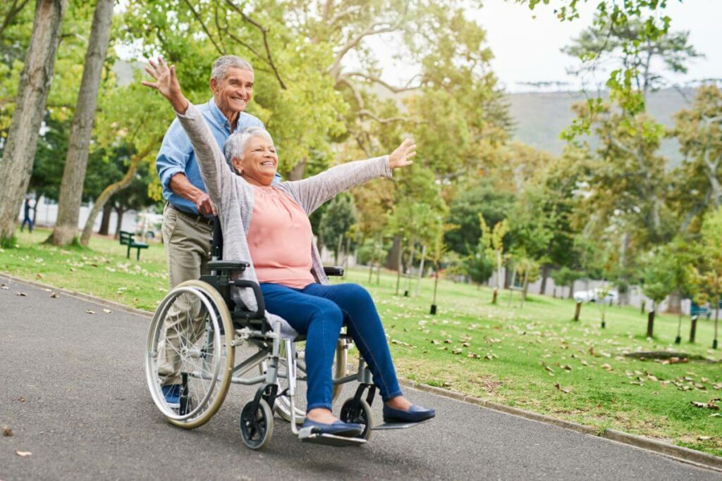 a senior man pushes his disabled wife in her wheelchair as they joyfully take a walk through the park dressed in adaptive clothing.