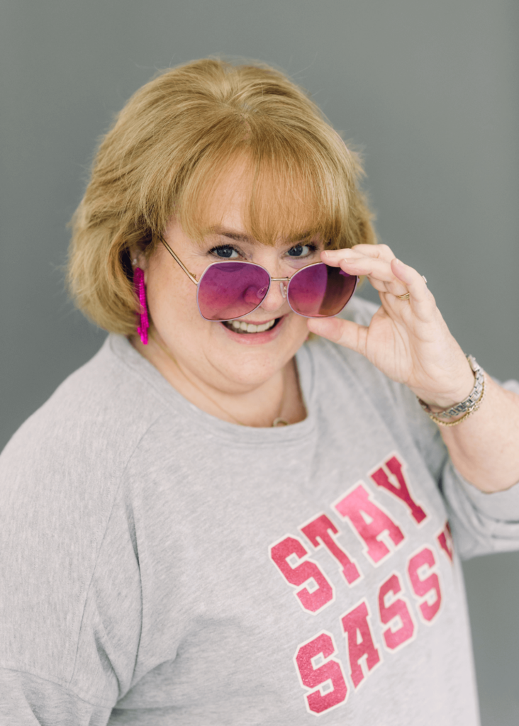 Author wearing a Stay Sassy sweatshirt smiling over a pair of sunglasses - Contact Page