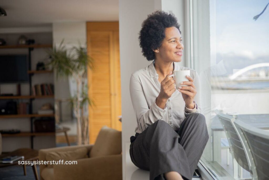 a middle age black woman sits on a window sill looking out a window as she relaxes and meditates.