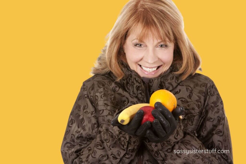 a middle age woman dressed in her winter coat and gloves holds three pieces of fruit for healthy snacks this winter