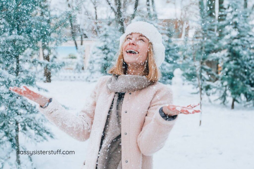 a middle aged woman stands in a snowstorm with her hands open facing the sky and a big smile on her face