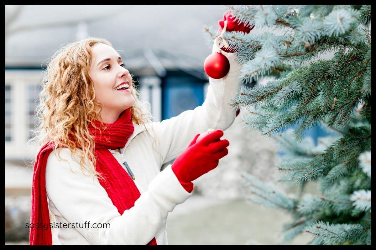 a festive woman hangs a red christmas tree ornament on a tree outside her house after reading about holiday lifestyle tips that recommend getting outside during the winter