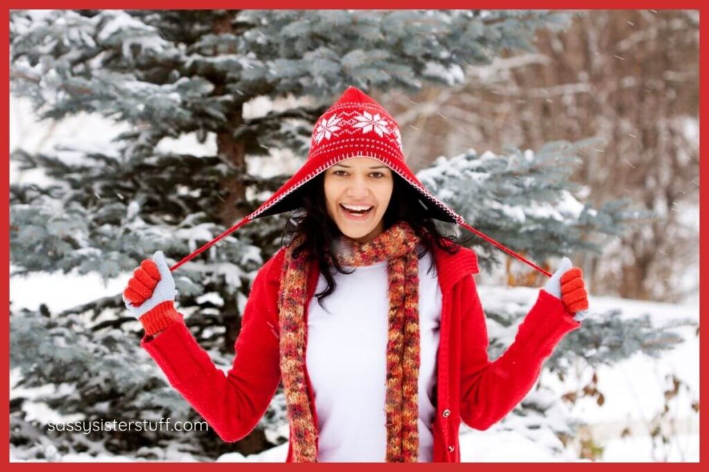 a festive woman dressed in red gloves, a red hat, and a red coat stands in the snow and smiles at the camera