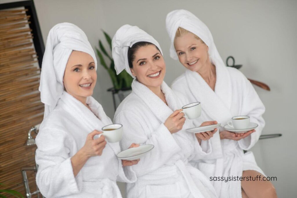 three middle aged women dressed in white robes and head wraps practice mindful aging by engaging in self-care at a spa.