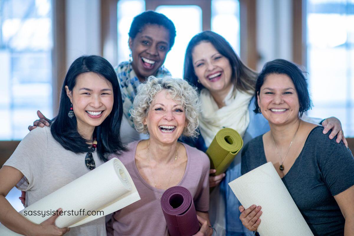 five happy middle aged woman in a yoga studio holding their rolled up mats smiling for a camera