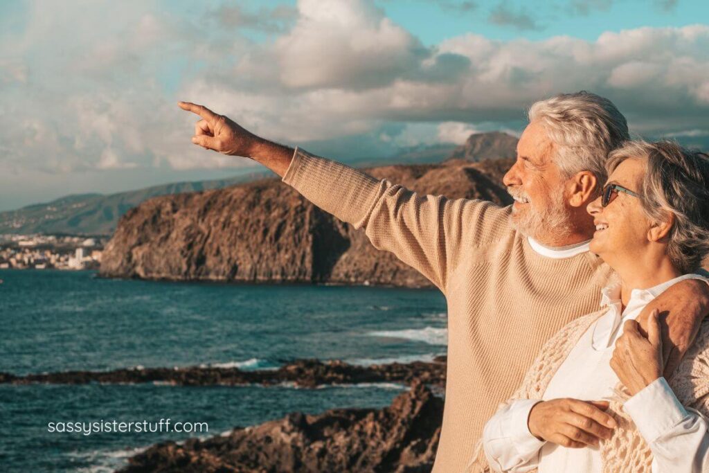 a retired couple stand on a rock cliff overlooking a beautiful body of water as they enjoy adventures together