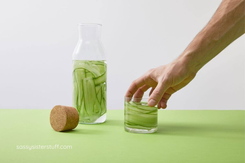 close up of a hand reaching for a glass of water that sits next to a pitcher of water with cucumber