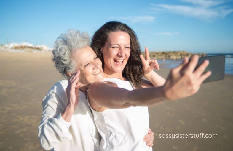 an adult woman and her aging mother enjoy taking selfies on the beach.