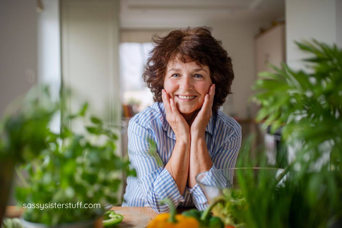 a midle age woman surrounds herself with beautiful indoor green plants and smiles at the camera.