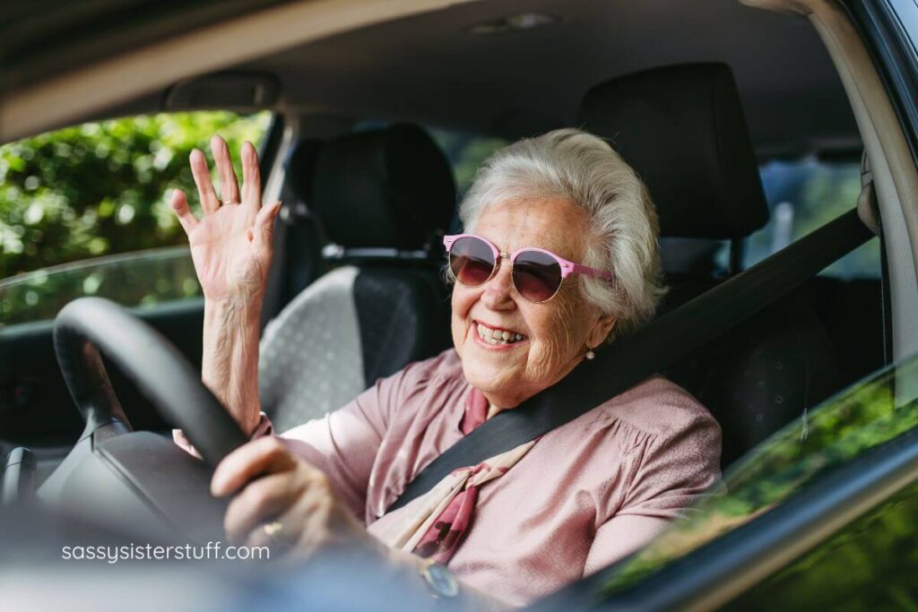 an elderly woman drives her car and waves at a neighbor.