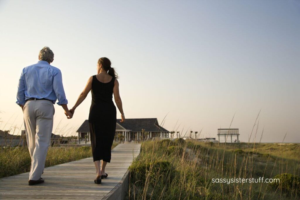 a middle aged couple walks hand in hand on a boardwalk over a marsh.