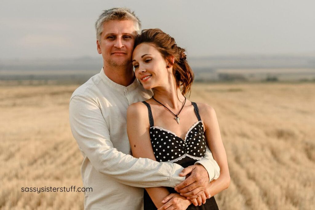 a man holds his wife affectionately on a beach as she rests her head on his cheek.