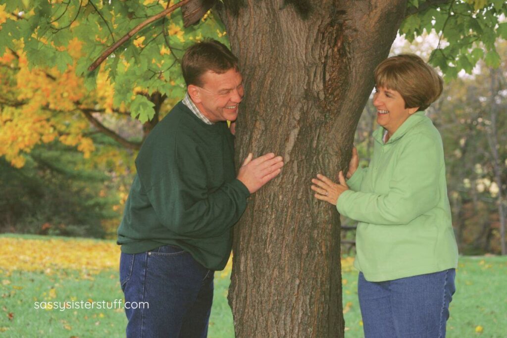 middle aged couple peeks around a large tree at each other.