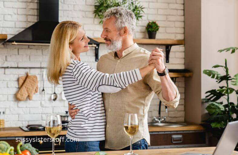 a midlife husband and wife dance in the kitchen and enjoy glasses of wine together.