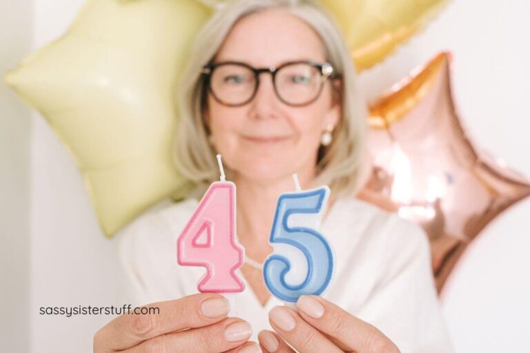 middle aged woman with three balloons behind her holds two candles for her 45th birthday.