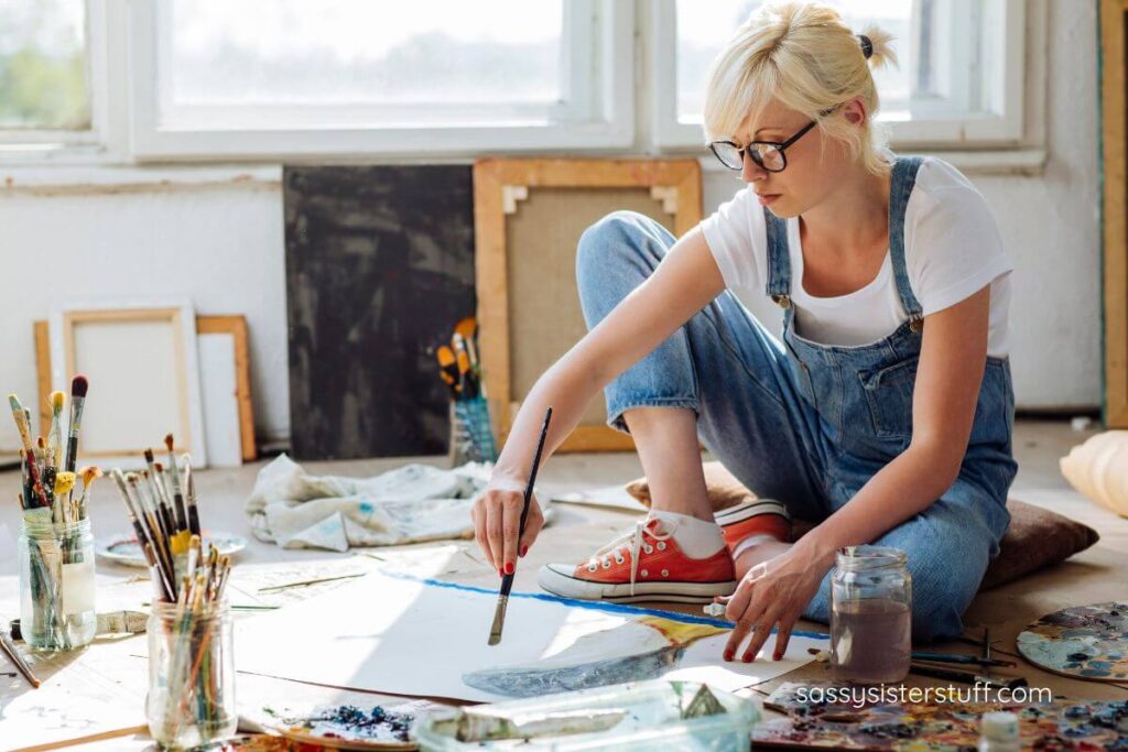 woman sitting on the floor with paint supplies around her, painting on a large canvas.