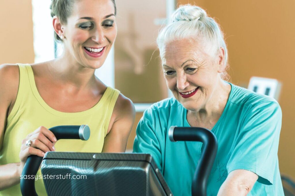 adult woman stands next to her aging mother and teaches her how to ride a stationary bike.