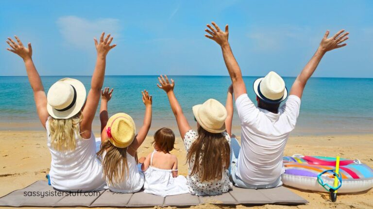 family sitting on the beach with hands in the air showing why traveling is important to happiness.