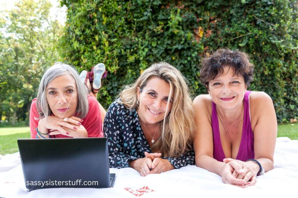 three middle aged females outside in a park laying on their bellies with a laptop.